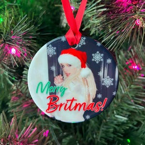 May include: A round Christmas ornament with a white background and a black and white image of a woman wearing a red Santa hat and a white sweater. The text "Merry Britmas!" is written in green and red letters on the ornament.