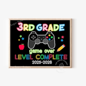 May include: A framed black sign with the words "3RD GRADE" in colorful letters, a gray video game controller, and the text "game over LEVEL COMPLETE 2025-2026". Includes a red apple, stars, and a yellow pencil.