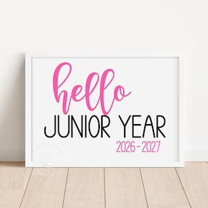 May include: A framed print with the words "hello JUNIOR YEAR" in pink and black, with the year range 2026-2027. The print is in a white frame, set against a white wall and light wood floor.