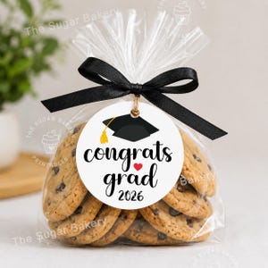 May include: A clear cellophane bag tied with a black ribbon, filled with chocolate chip cookies. A round tag with a graduation cap graphic and the words "congrats grad 2026" is attached. The Sugar Bakery logo is visible.