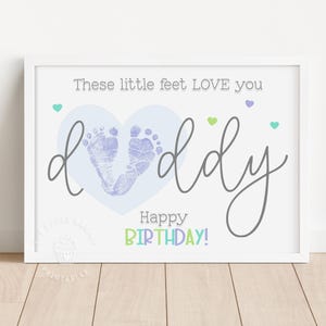Puede incluir: Una impresión enmarcada con las palabras "These little feet LOVE you daddy" en escritura gris. La palabra "daddy" está escrita sobre un corazón azul claro con dos huellas de bebé. Las palabras "Happy BIRTHDAY!" están en verde y azul.