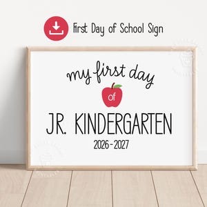May include: A rectangular sign in a light wood frame featuring the black text 'my first day of', a red apple illustration, the word 'of' in white, and the black text 'JR. KINDERGARTEN' followed by the school year '2026-2027' on a white background.