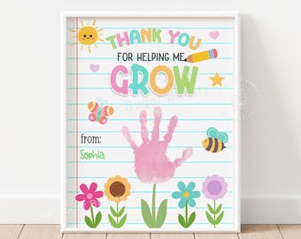Può includere: Una stampa incorniciata con un bordo bianco presenta il testo "Thank You For Helping Me Grow" in lettere colorate. Un'impronta di mano rosa è al centro, con illustrazioni di un sole, fiori, una farfalla e un'ape. Il nome "Sophia" è scritto sotto.