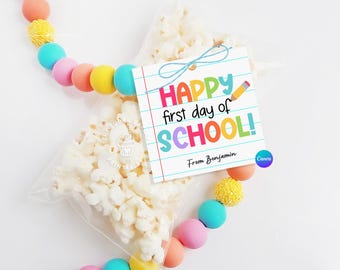 Könnte beinhalten: Ein farbenfroher, druckbarer Geschenkanhänger mit dem Text "Happy first day of SCHOOL!" und "From Benjamin". Der Anhänger ist mit einem Bleistift und einem blauen Band verziert. Der Anhänger liegt auf einem Beutel Popcorn.