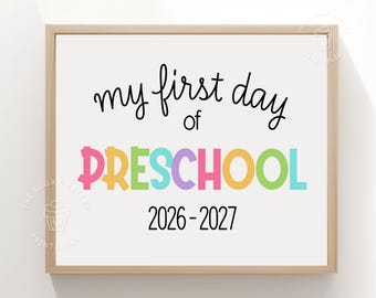 May include: A rectangular wooden frame containing a white sign with black text that reads my first day of, followed by the word PRESCHOOL in multicolored letters, and the years 2026-2027 in black text at the bottom of the sign.