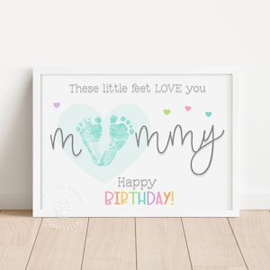 May include: A framed print with the text "These little feet LOVE you mommy" and "Happy BIRTHDAY!". The artwork features two teal-colored baby footprints inside a light blue heart, with small colorful hearts above the word "mommy".