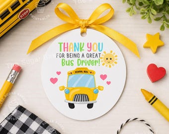 Puede incluir: Una etiqueta circular blanca con una cinta amarilla, con el texto "THANK YOU FOR BEING A GREAT Bus Driver!". La etiqueta incluye una ilustración de autobús escolar amarillo, corazones y un sol sonriente. Un autobús escolar de juguete amarillo está en el fondo.