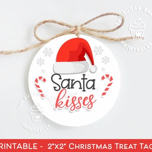Può includere: Un'etichetta rotonda per dolcetti natalizi con un cappello rosso di Babbo Natale, bastoncini di zucchero e le parole "Santa kisses". L'etichetta bianca ha un cordino marrone per appenderla. Il testo "The Sugar Bakery Printables" è presente.