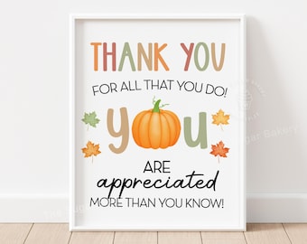 Puede incluir: Una impresión enmarcada con las palabras "THANK YOU" en naranja y rosa, y "YOU" en verde y marrón. Una calabaza naranja y hojas de otoño decoran la impresión. El texto "ARE APPRECIATED MORE THAN YOU KNOW!" está en negro.