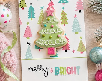 Op de afbeelding: Een rechthoekige wenskaart met een patroon van kleurrijke kerstbomen in tinten roze, groen en blauw. Een versierd kerstboomkoekje met groen glazuur, witte krullen en een roze ster ligt op de kaart boven de tekst merry & BRIGHT.