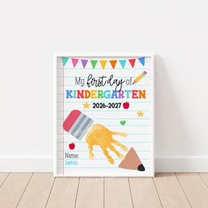 May include: A white poster with blue horizontal lines featuring the text My first day of KINDERGARTEN 2026-2027. It includes a colorful bunting, a yellow pencil, red apples, yellow stars, a green heart, and a yellow handprint shaped like a pencil.