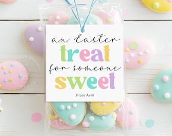 Könnte beinhalten: Ein weißes Geschenkanhänger mit dem Text "an Easter treat for someone sweet" in bunten Buchstaben. Der Anhänger ist mit einem blauen Band gebunden und liegt auf einem durchsichtigen Plastikbeutel mit pastellfarbenen Keksen in Form von Ostereiern.