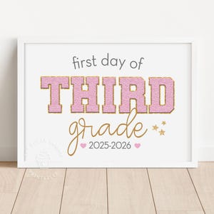 May include: A framed print with the text "first day of THIRD grade" in pink and gold lettering. The words "grade" and "2025-2026" are in gold script. Small gold stars and pink hearts accent the design. The print is in a white frame.