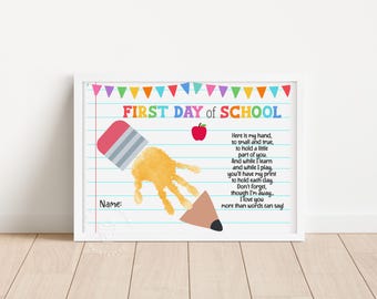 May include: A white-framed print featuring a lined paper background with colorful bunting at the top. It displays the text FIRST DAY OF SCHOOL, a red apple, a yellow handprint shaped like a pencil, and a poem about holding a handprint.