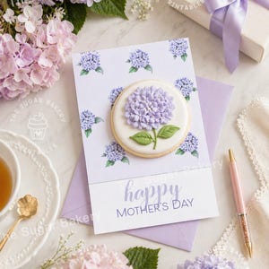 Puede incluir: Una tarjeta del Día de la Madre con una galleta de hortensia, un sobre y una caja de regalo. La tarjeta dice "happy MOTHER'S DAY" en escritura lavanda. La galleta está decorada con una flor de hortensia morada. La escena incluye flores, un bolígrafo y una taza de té.
