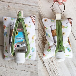May include: White zippered pouch with a floral pattern and a small bottle of Germ-X hand sanitizer inside. The pouch has a green zipper and a silver key ring.