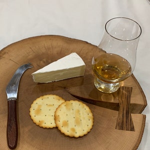 May include: A wooden serving board with a wedge of brie cheese, two crackers, a glass of amber liquid, and a cheese knife. The board is made from a cross-section of a tree trunk.