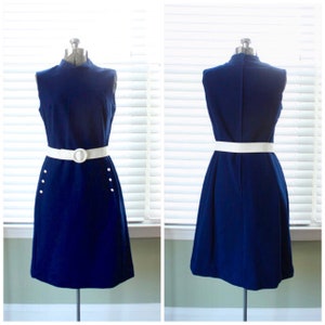 May include: A sleeveless navy blue dress with a high neckline and a white belt. The dress features white buttons on the front and is displayed on a mannequin. The dress is a vintage style.