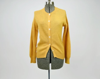 Pode incluir: Um cardigan amarelo mostarda com mangas compridas e decote redondo. O suéter abotoado é exibido em um manequim. O suéter é feito de um material macio e tricotado.