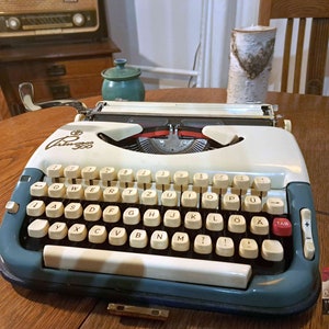 Puede incluir: Una máquina de escribir vintage blanca y azul con un teclado negro y la palabra "Olympia" impresa en la parte delantera. La máquina de escribir está sobre una mesa de madera.