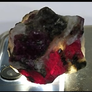 Può includere: Una gemma grezza con una miscela di colori. La pietra presenta un colore rosso intenso in alcune aree, a contrasto con macchie nere, bianche e gialle. La forma generale è irregolare, mostrando la sua forma naturale.