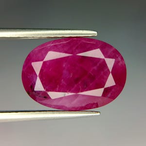 May include: An oval-cut, vibrant magenta-red gemstone featuring a distinct six-pointed star pattern across its faceted surface. The stone is held horizontally by two metallic prongs, showcasing its translucent quality and deep, saturated colour.