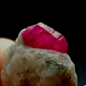 以下が含まれることがあります： 原石のルビー結晶のクローズアップ。ルビーは鮮やかで深いピンク色で、ファセット加工された表面を持っています。明るいグレーと白の石の母岩に、濃い斑点とともにセットされています。