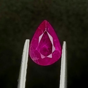 Può includere: Una gemma a forma di pera di un vibrante colore rosa magenta, tenuta da una pinzetta in metallo color argento. La pietra sfaccettata mostra caratteristiche di purezza interna e una parte superiore appuntita.