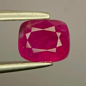 Könnte beinhalten: Ein kissenförmig geschliffener, leuchtend pink-roter Edelstein, der von einer Metallpinzette gehalten wird. Der Stein weist ein deutliches, helleres, sternförmiges Einschlussmuster auf, das in seiner facettierten Oberfläche sichtbar ist.
