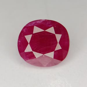 May include: A faceted, oval-shaped ruby gemstone. The deep red stone features a starburst pattern of white facets in the center. The gem is set against a plain white background.