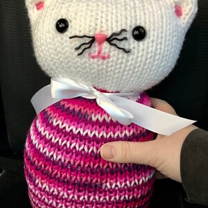 May include: White crocheted cat with pink nose and black whiskers, wearing a pink and white striped sweater with a white ribbon around its neck.