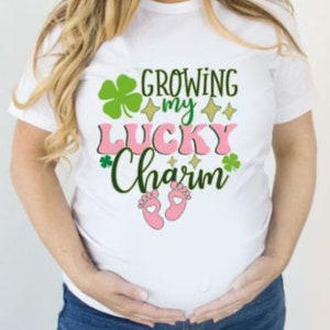 May include: White t-shirt with the text "Growing my Lucky Charm" in green and pink lettering. There are shamrock and star graphics around the text.  Pink baby footprints are below the text.