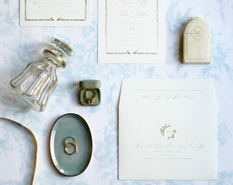 May include: An elegant wedding invitation suite featuring a cream-colored invitation, RSVP card, and envelope with gold accents. Includes a glass perfume bottle, a ring in a green box, and two gold rings on a gray oval dish. The background is a floral pattern.