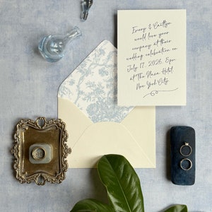 Peut inclure: Une enveloppe blanche avec une doublure à motif floral bleu. L'enveloppe est adressée à Emery et Caitlyn pour leur célébration de mariage le 17 juillet 2026 à 17h00 au Plaza Hotel de New York.