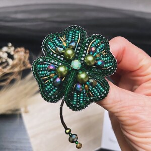 Op de afbeelding: Een broche in de vorm van een vierbladig klavertjevier, gemaakt met groene en gouden kralen. Het klavertjevier is versierd met groene en iriserende kralen.