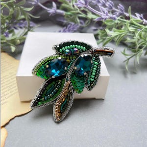 May include: A handcrafted leaf-shaped brooch with intricate beadwork. The brooch features green sequins, beads, and two large, teal-colored gemstones. The design includes silver-colored metal accents and a brown stem, creating a nature-inspired accessory.