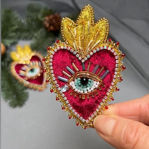 Può includere: Un broche a forma di cuore in velluto rosso con un occhio di perline e dettagli dorati. L'occhio è circondato da perline dorate e argentate e ha un'iride verde e una pupilla nera.