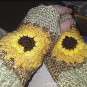 May include: A pair of handmade crochet fingerless gloves. The gloves feature a sunflower design with yellow petals and a brown center. The base of the gloves is a mix of green, brown, and tan yarn.