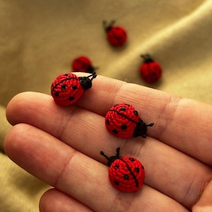 May include: Several small, handcrafted crochet ladybugs featuring bright red bodies with black spots and black heads. Three of these miniature insect figures are positioned on a palm, while two additional ladybugs are placed in the background.