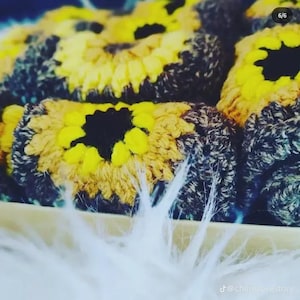 May include: Close-up of several crocheted sunflower-shaped items. Each piece features a yellow petal design around a black center, with a brown and gray border. The items are arranged in a wooden tray, with white fluffy material in the foreground.