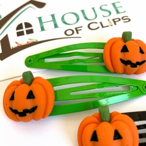 Op de afbeelding: Drie groene plastic haarclips met oranje pompoen-vormige bedels. Elke pompoen heeft een jack-o'-lantern gezicht met zwarte ogen, een neus en een mond.