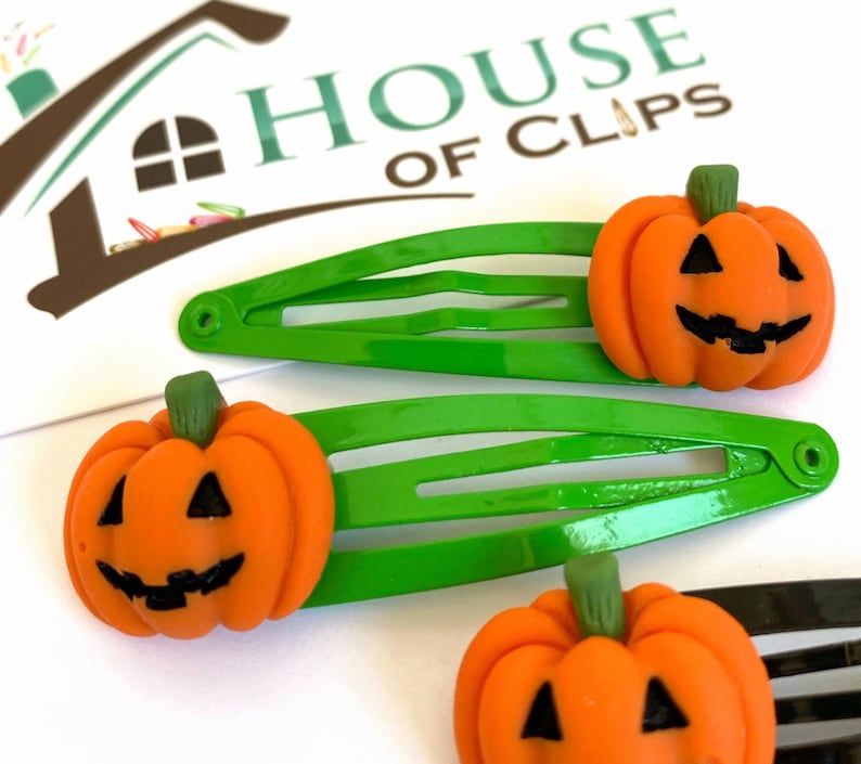 Op de afbeelding: Drie groene plastic haarclips met oranje pompoen-vormige bedels. Elke pompoen heeft een jack-o'-lantern gezicht met zwarte ogen, een neus en een mond.