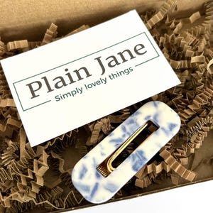 Op de afbeelding: Een rechthoekige haarclip met een goudkleurige sluiting. De clip heeft een witte basis met een blauw patroon. De clip ligt in een kartonnen doos met bruine papieren vulling. Een witte kaart met de tekst "Plain Jane Simply lovely things" staat ook in de doos.
