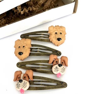 May include: Four bronze-toned metal hair clips with dog-shaped embellishments. Two clips feature light brown dogs, while the other two have brown and white dogs with pink tongues. The box in the background has the text "HOUSE OF CLIPS".