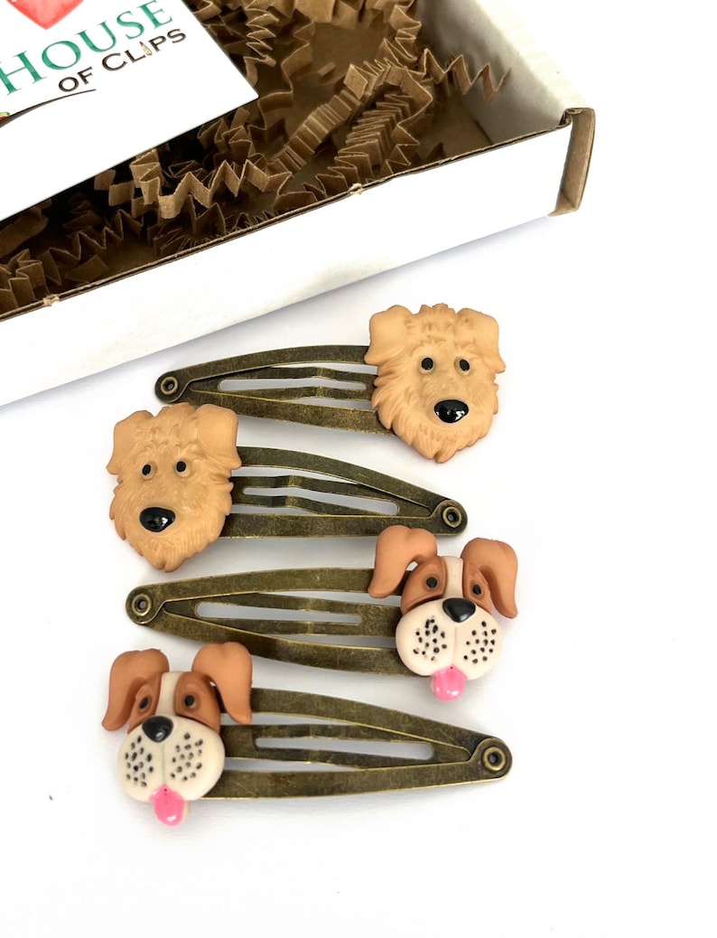 May include: Four bronze-toned metal hair clips with dog-shaped embellishments. Two clips feature light brown dogs, while the other two have brown and white dogs with pink tongues. The box in the background has the text "HOUSE OF CLIPS".