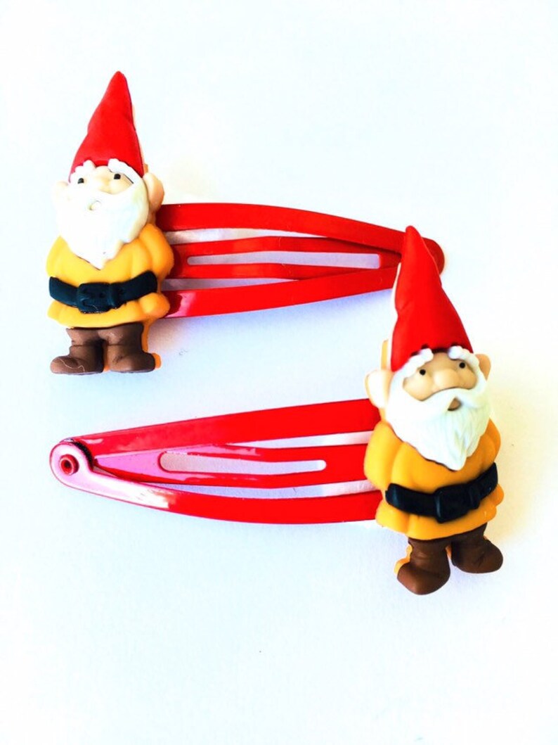 May include: Two red hair clips with a gnome design. Each gnome is wearing a yellow shirt, brown pants, and a red hat.