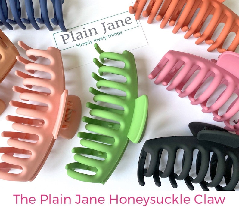 Puede incluir: Un conjunto de cuatro pinzas para el pelo en diferentes colores: rosa, verde, negro y naranja. Las pinzas tienen todas el mismo estilo, con una forma curva y varios dientes. El texto "Plain Jane - Simply lovely things" es visible en la imagen.