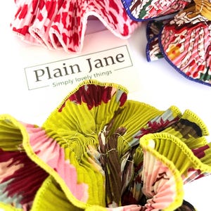 May include: Three fabric hair scrunchies in a variety of floral patterns. The scrunchies are made of pleated fabric with a contrasting trim. The scrunchies are arranged on a white surface with a business card that reads "Plain Jane - Simply lovely things".