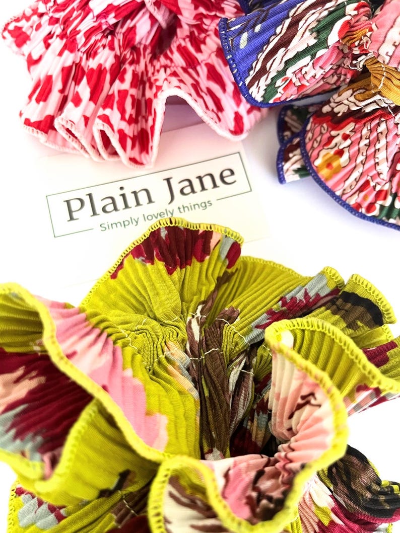 May include: Three fabric hair scrunchies in a variety of floral patterns. The scrunchies are made of pleated fabric with a contrasting trim. The scrunchies are arranged on a white surface with a business card that reads "Plain Jane - Simply lovely things".