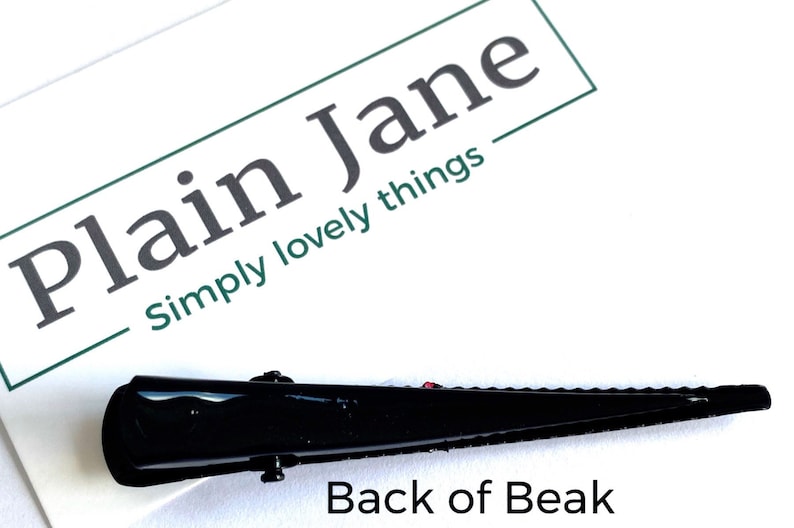 May include: A black hair clip with a beak-like design. The clip is glossy and has a textured interior for grip. The image includes the text "Plain Jane Simply lovely things" and "Back of Beak".